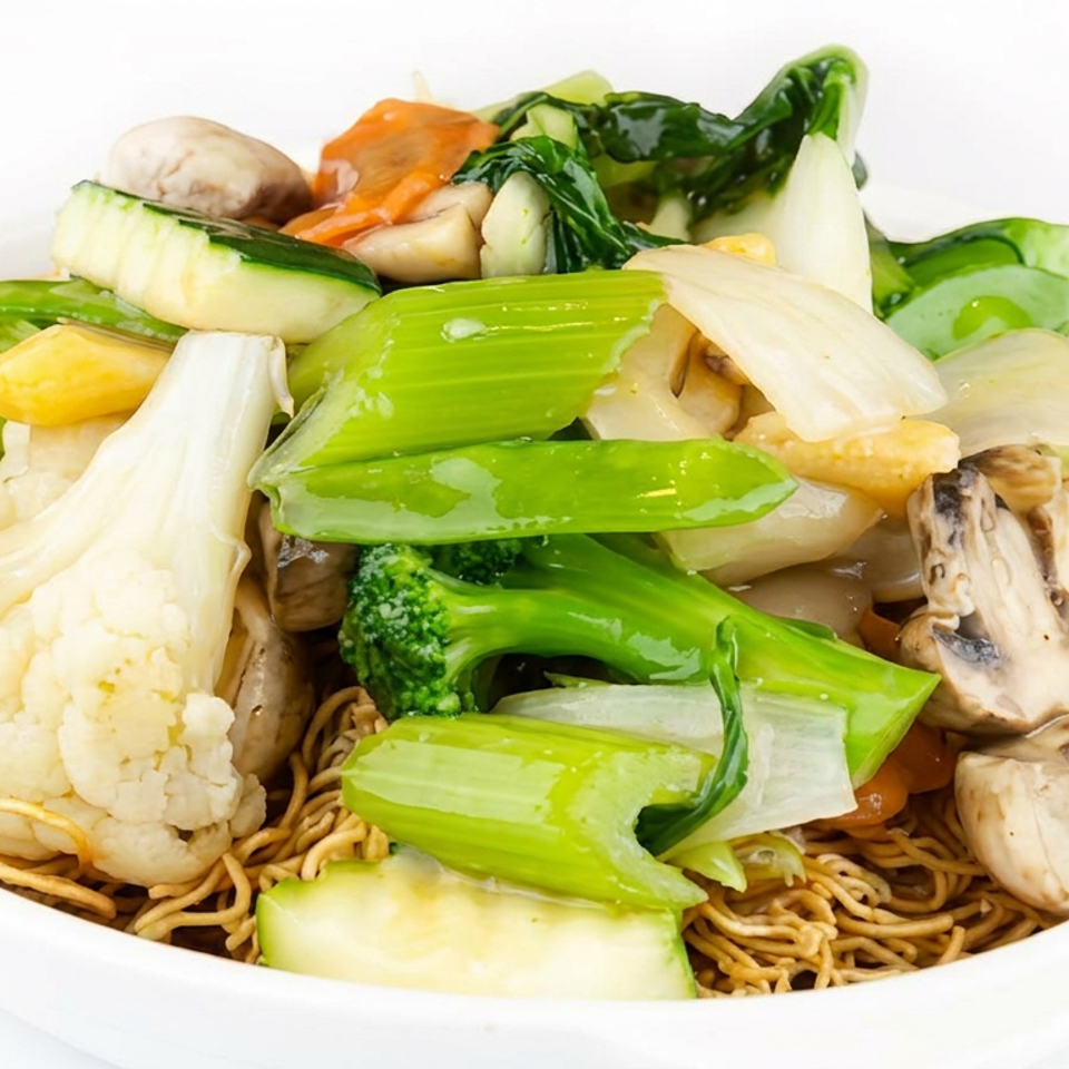 Crispy noodles topped with stir-fried mixed vegetables, mushrooms, cauliflower, broccoli, bok choy, and zucchini in a white bowl.
